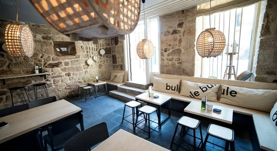Ambiente acogedor para comer en Allariz. Mesas de madera, bancos con cojines y lámparas de mimbre en un local de piedra.