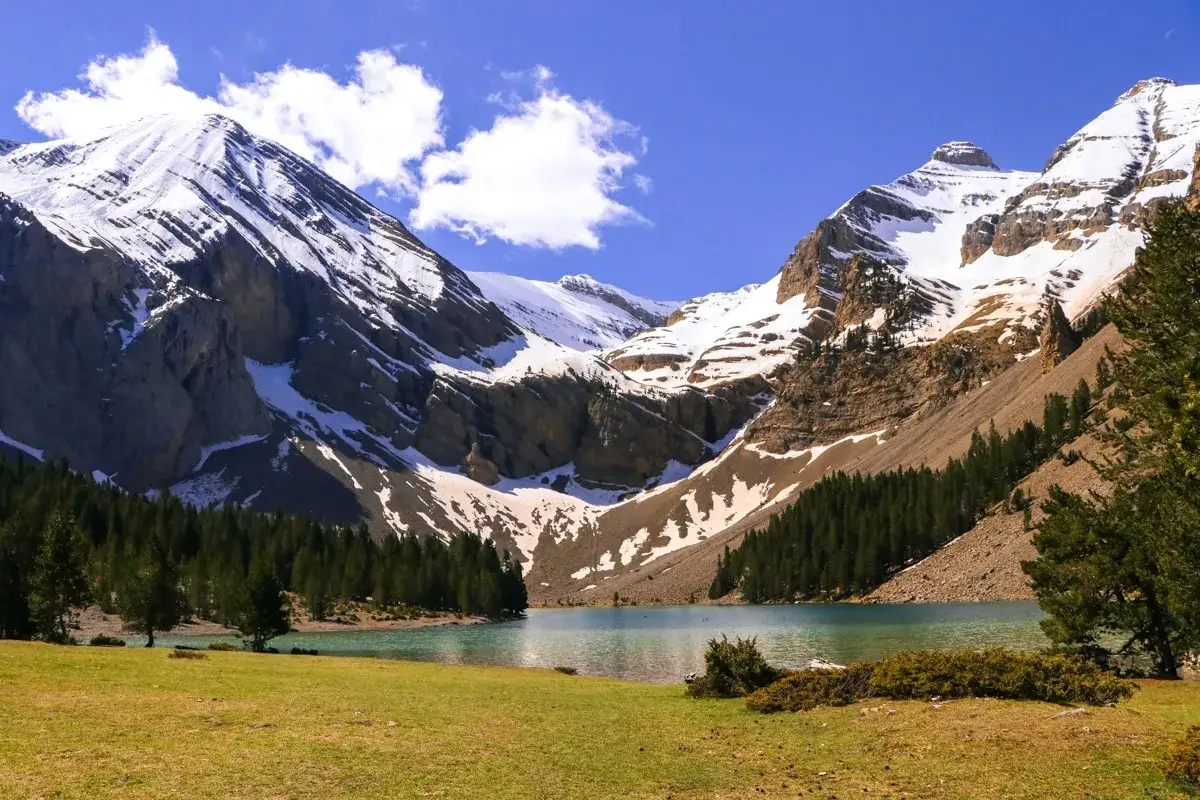 Majestuosas montañas nevadas rodean el ibon de Plan, un lago turquesa enmarcado por pinos y prados verdes.