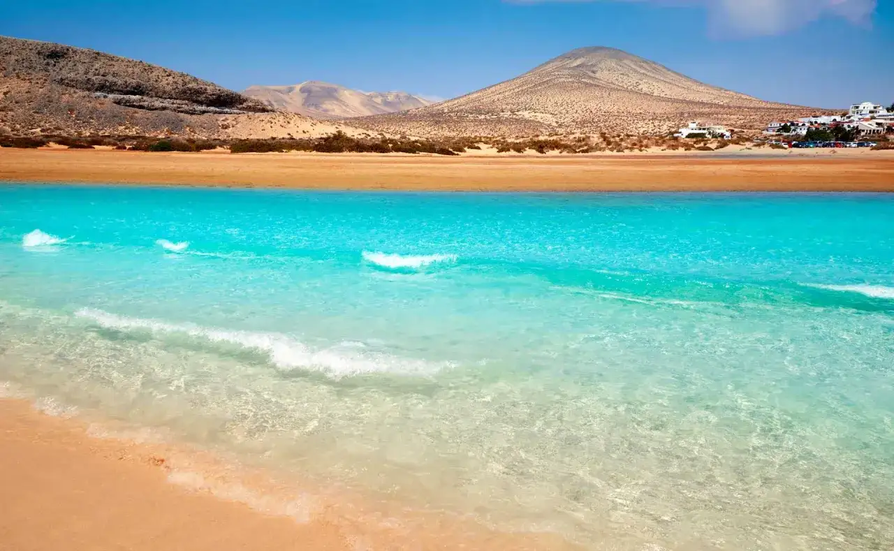 Aguas turquesas y arena dorada en las playas Fuerteventura, con montañas áridas y casas blancas al fondo.