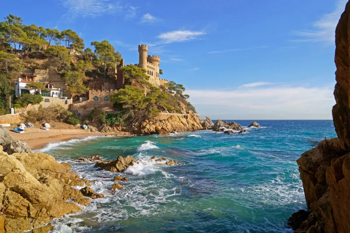 Castillo medieval sobre acantilados rocosos en Cala Bona, Barcelona. Mar azul turquesa y cielo despejado.