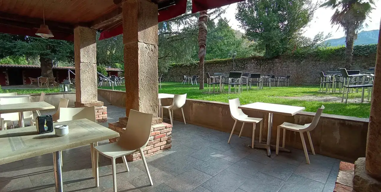 Terraza de restaurante con mesas y sillas, ideal para saber donde comer en Lierganes. Jardín verde y muro de piedra al fondo.