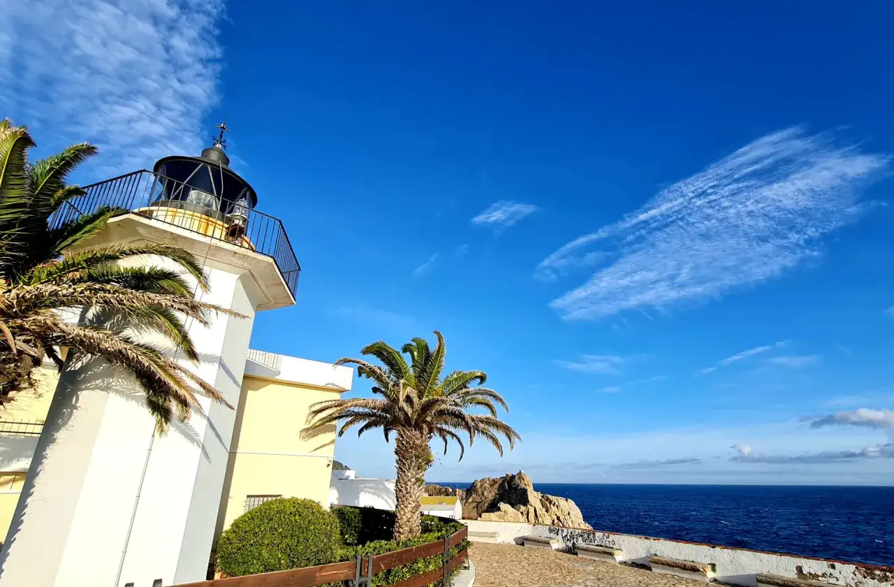 El faro de Palamós se alza imponente bajo un cielo azul intenso, flanqueado por palmeras y con el mar Mediterráneo de fondo.