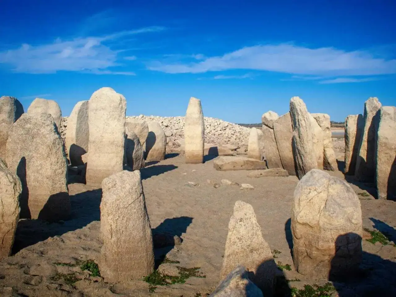 Un c&iacute;rculo de grandes piedras verticales, que es un cromlech, se alza sobre un terreno arenoso bajo un cielo azul brillante.
