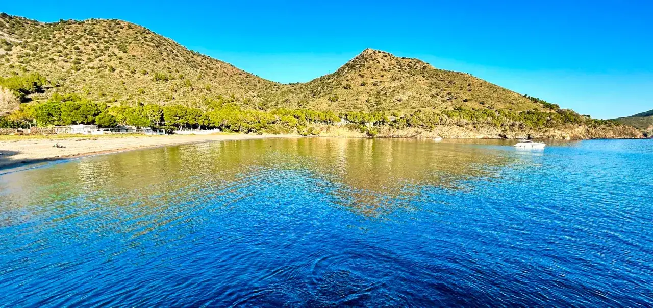 Aguas cristalinas reflejan las colinas verdes y el cielo azul en la cala Joncols plage. Una pequeña embarcación descansa en la bahía.