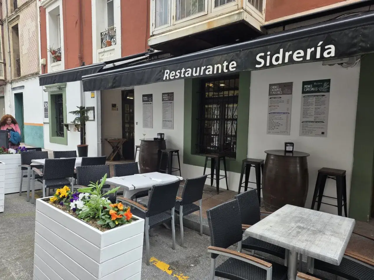 Terraza de restaurantes en Llanes con mesas y sillas negras, barriles de sidra y maceteros con flores.