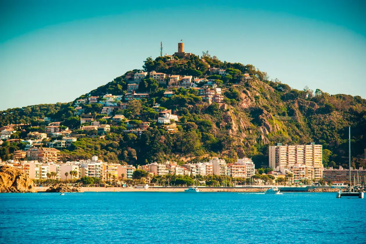 Vista de la costa con casas en la ladera y el castillo de Blanes en la cima. El mar azul y los barcos completan la escena.