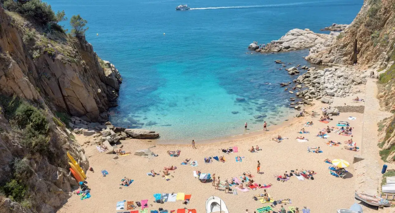 Gente disfrutando del sol en la arena dorada de la cala Tossa de Mar, con aguas cristalinas y rocas escarpadas.