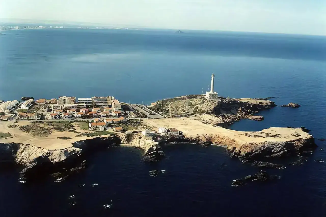 Vista aérea de un pueblo costero con un faro blanco en un promontorio rocoso. El mar azul profundo rodea la cala roja y las casas blancas.