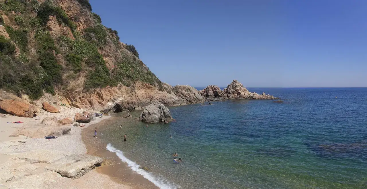 Les platges de Blanes ofereixen aigües cristal·lines i roques imponents sota un cel blau. Gent gaudeix del sol i del mar.