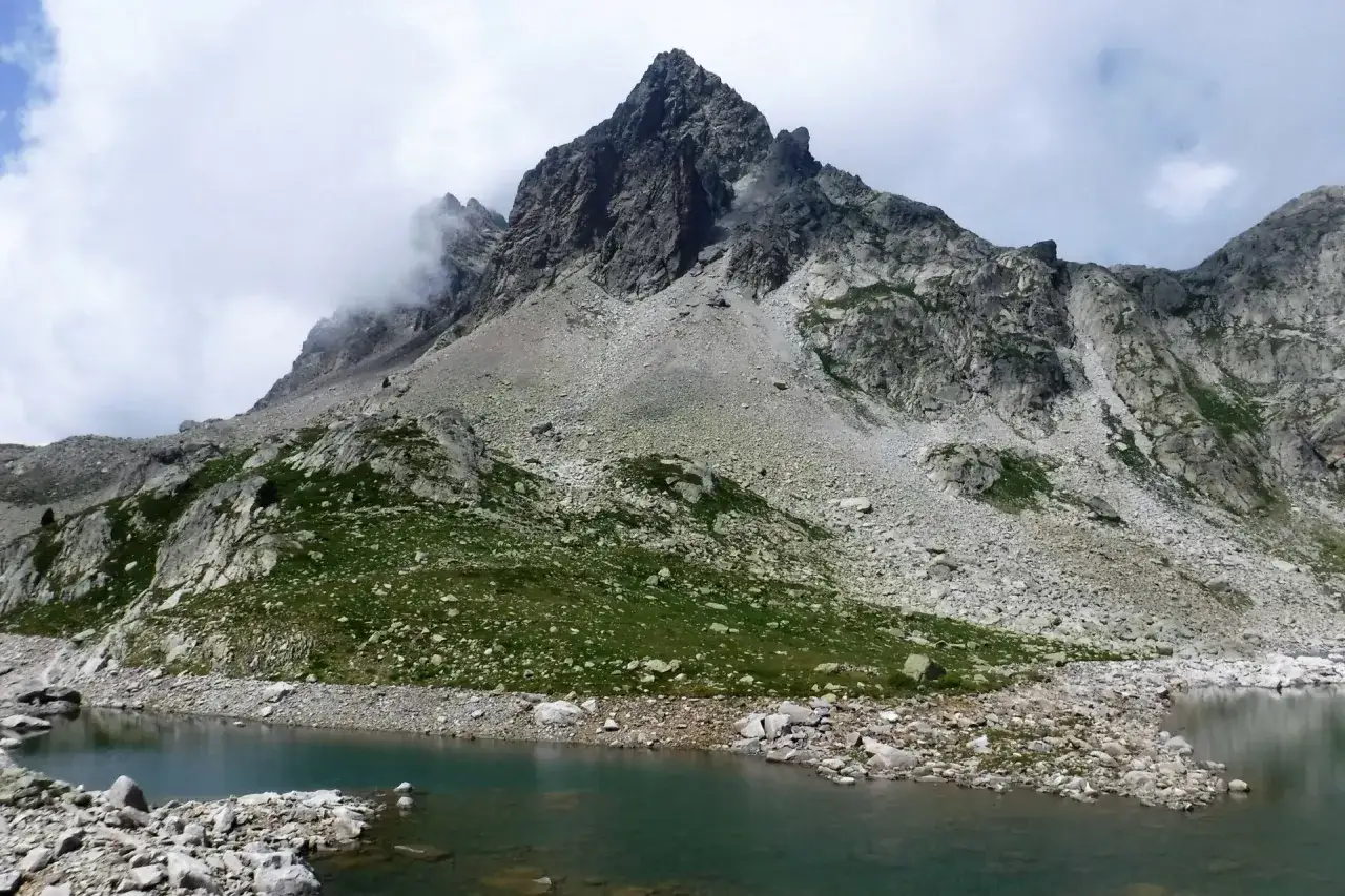 Majestuoso pico rocoso coronado por nubes, con un lago de aguas tranquilas y vegetación verde en primer plano. Los ibones de Arriel reflejan la imponente montaña.