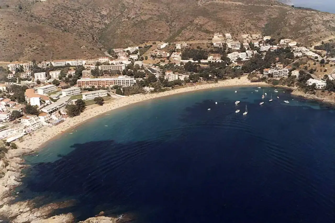 Vista aérea de la playa Almadrava en Roses, con edificios blancos y barcos anclados en aguas turquesas.