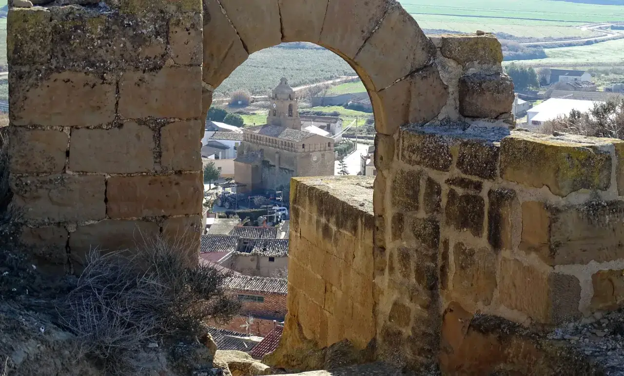 Vista de un pueblo a trav&eacute;s de un arco de piedra. Se ve una iglesia con torre y tejados de teja. El paisaje circundante es verde y rural, con campos y vegetaci&oacute;n. La alberuela de tubo se integra en la arquitectura antigua.