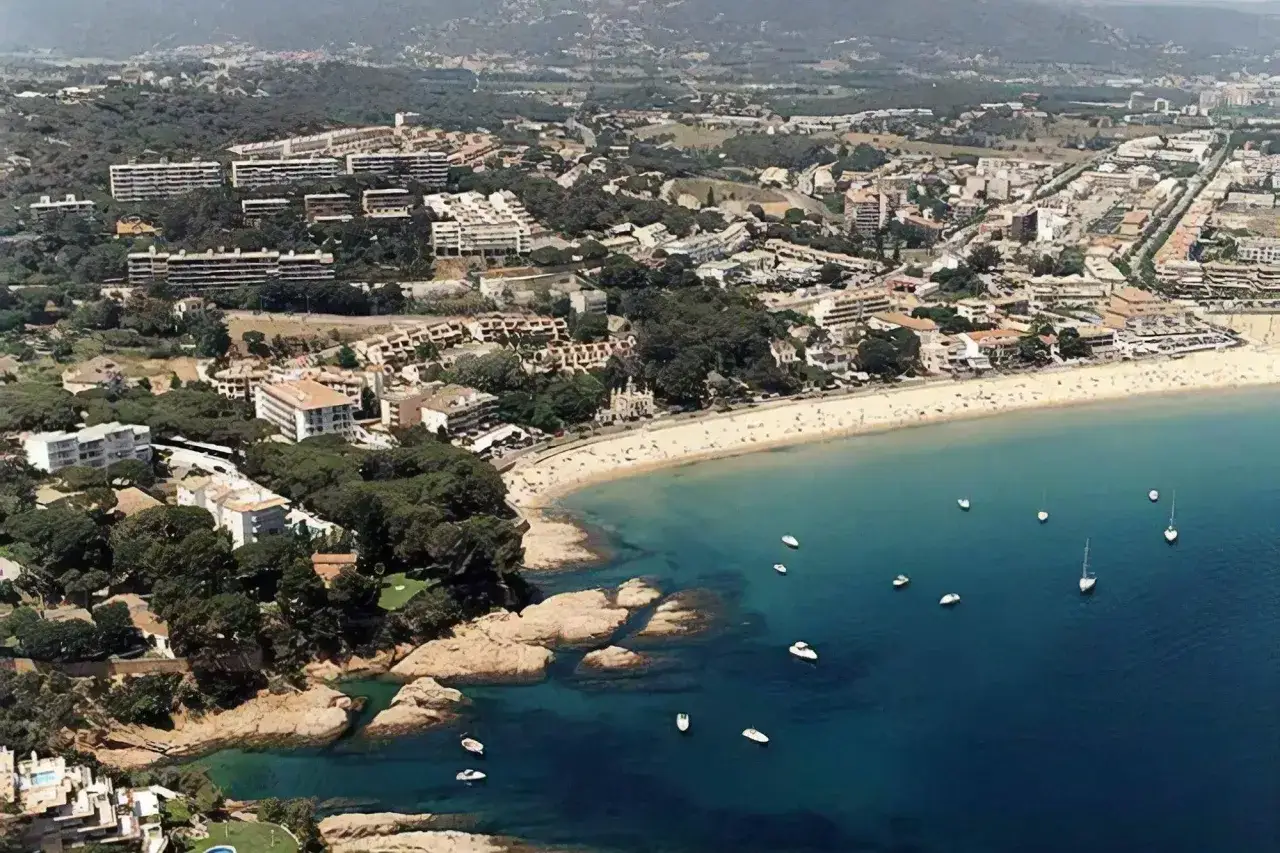 Vista a&eacute;rea de la platja Sant Pol en Sant Feliu de Gu&iacute;xols, con edificios, &aacute;rboles y barcos en el mar azul.