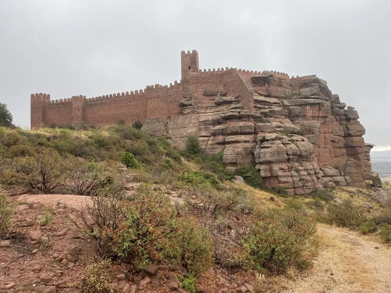Imponente castillo de Villar del Salz coronando una formaci&oacute;n rocosa rojiza, con vegetaci&oacute;n baja y un cielo nublado.