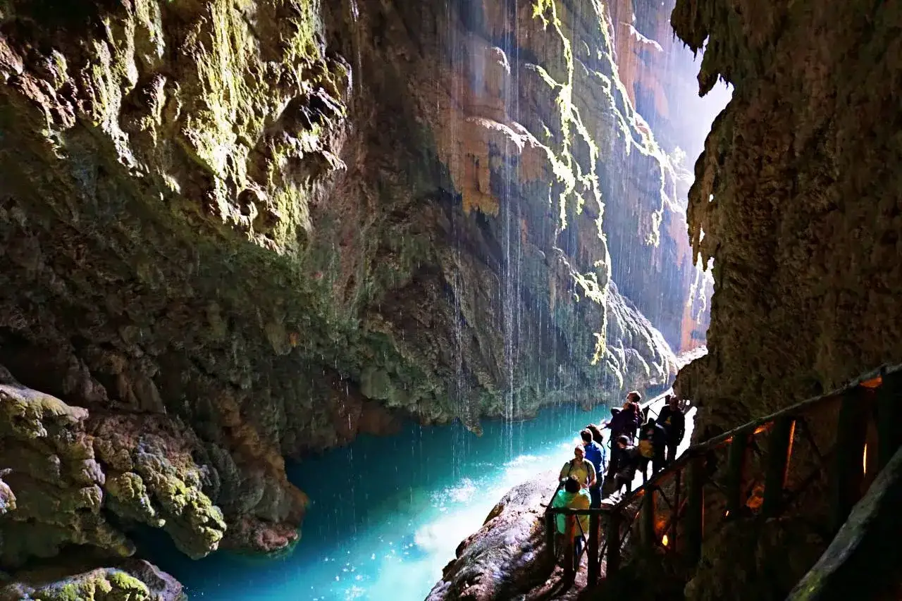 Turistas caminan por un sendero de madera junto a un río turquesa en el interior de una cueva, con cascadas cayendo de las rocas. Es como un monasterio de piedra en Zaragoza.