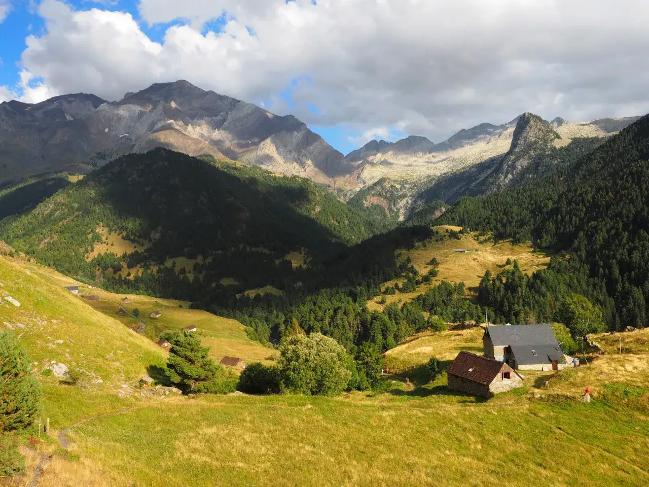 Paisaje montañoso con casas rurales, bosques y prados verdes. El sol ilumina la ruta de los tres refugios, invitando a la aventura.