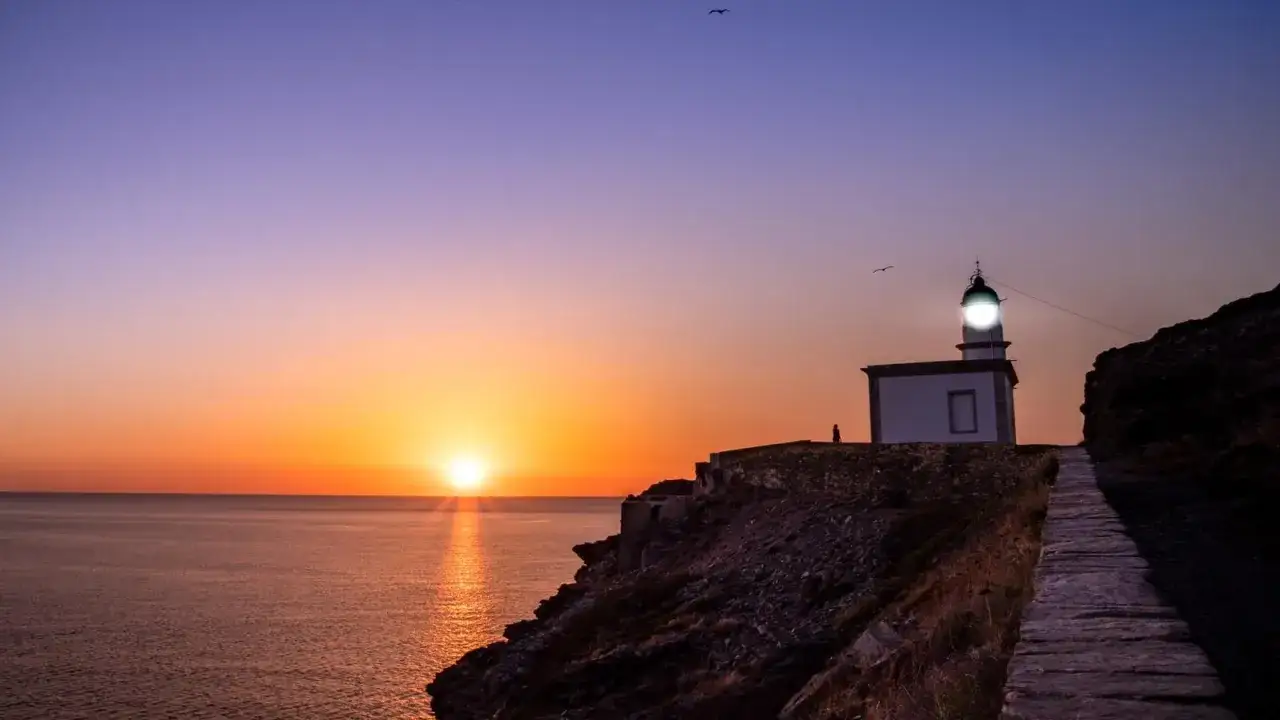 El faro de Cala Nans ilumina el atardecer sobre el mar. Una silueta solitaria camina por el sendero de piedra.