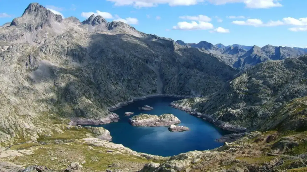 Los ibones de Bachimaña, un lago azul intenso rodeado de imponentes montañas rocosas y picos nevados bajo un cielo azul con nubes blancas.
