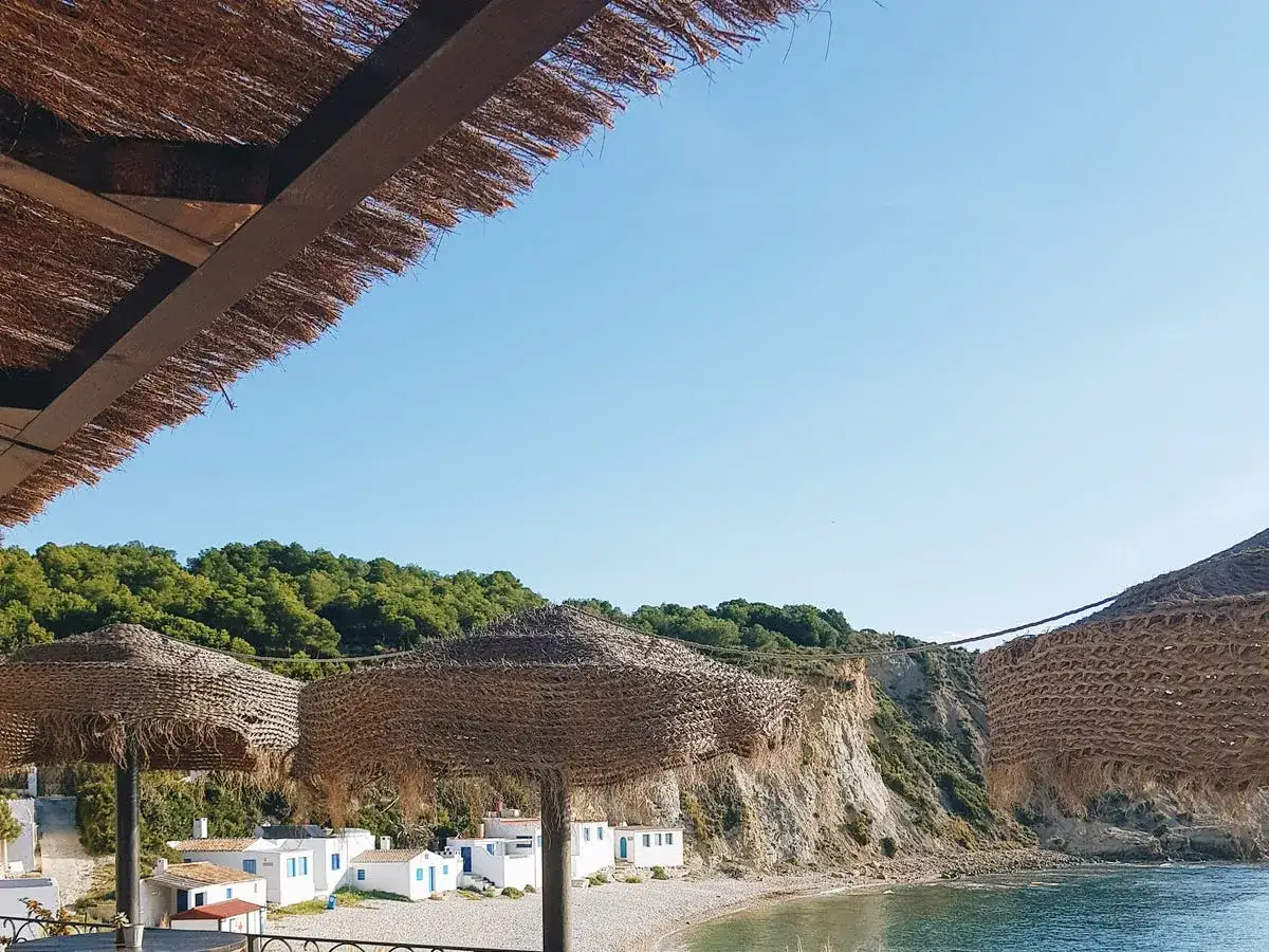 Sombrillas de paja cubren un rinc&oacute;n ideal donde comer en J&aacute;vea, con casitas blancas y acantilados verdes al fondo.
