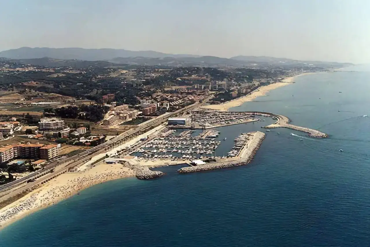 Vista aérea de la playa de les barques, con un puerto deportivo lleno de barcos, arena dorada y edificios costeros.