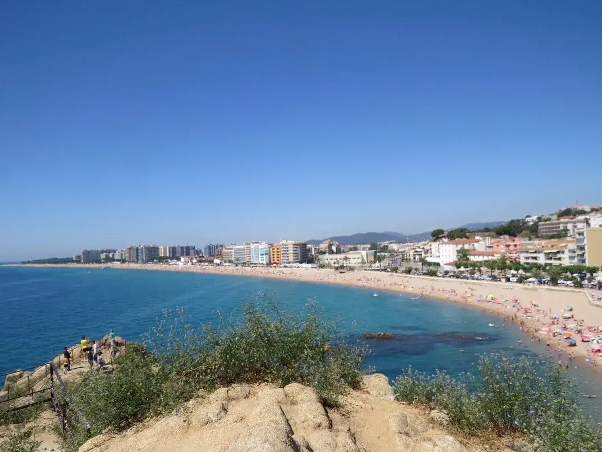 Vista panorámica de la playa S'Abanell en Blanes, con el mar azul, arena dorada y edificios costeros bajo un cielo despejado.
