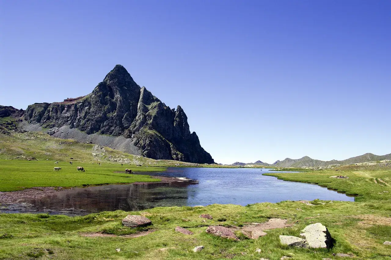 Majestuoso pico rocoso junto a un lago sereno en los ibones del Valle de Tena. Vacas pastan en la verde pradera.