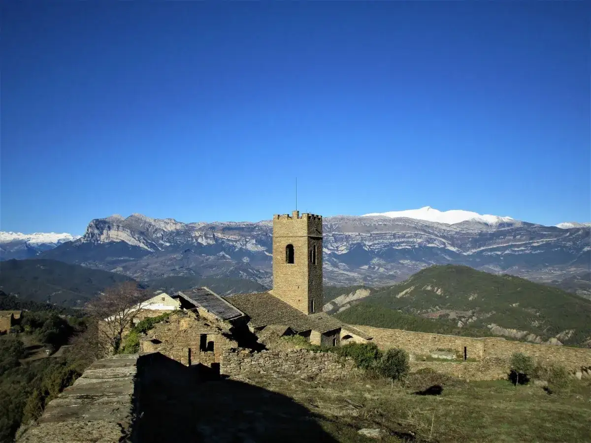 Antiguo muro de roda con torreón de piedra y ruinas, rodeado de montañas nevadas bajo un cielo azul intenso.