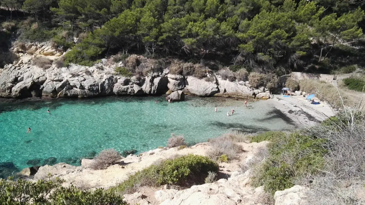 Es caló de la bella dona, una cala escondida con aguas turquesas y rocas escarpadas, donde algunas personas disfrutan del sol y el mar.