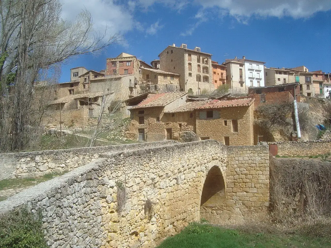 Un puente de piedra cruza un arroyo, llevando a un pueblo con casas de piedra y tejados rojos. Al fondo, se alza la torre de las arcas, vigilante del paisaje.