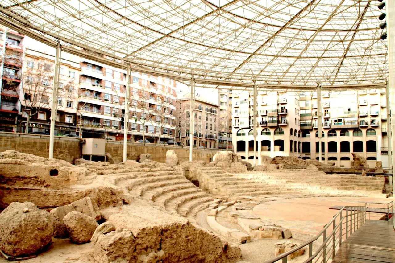 Antiguas ruinas romanas en Zaragoza, bajo una moderna cubierta metálica, con edificios modernos al fondo.