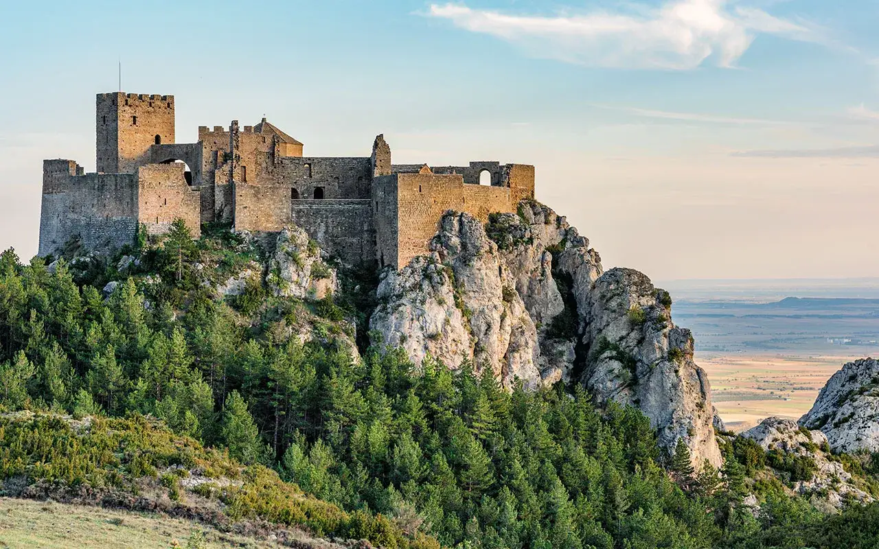 Castillo de Loarre, un lugar imprescindible que ver en Huesca, se alza majestuoso sobre un pe&ntilde;asco rocoso, rodeado de pinos y con vistas a un extenso paisaje.