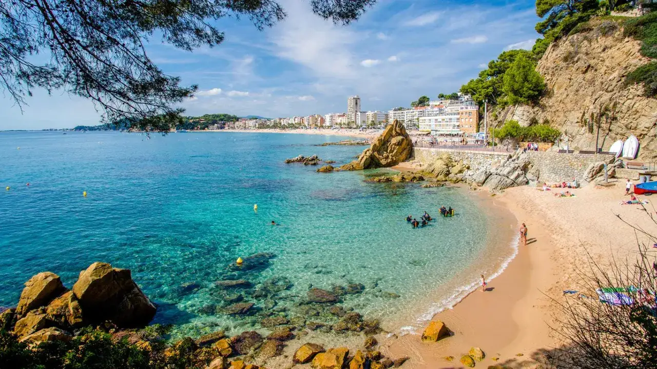 Una caleta en la Costa Brava con aguas cristalinas, buceadores explorando el fondo rocoso y una playa con gente disfrutando del sol.