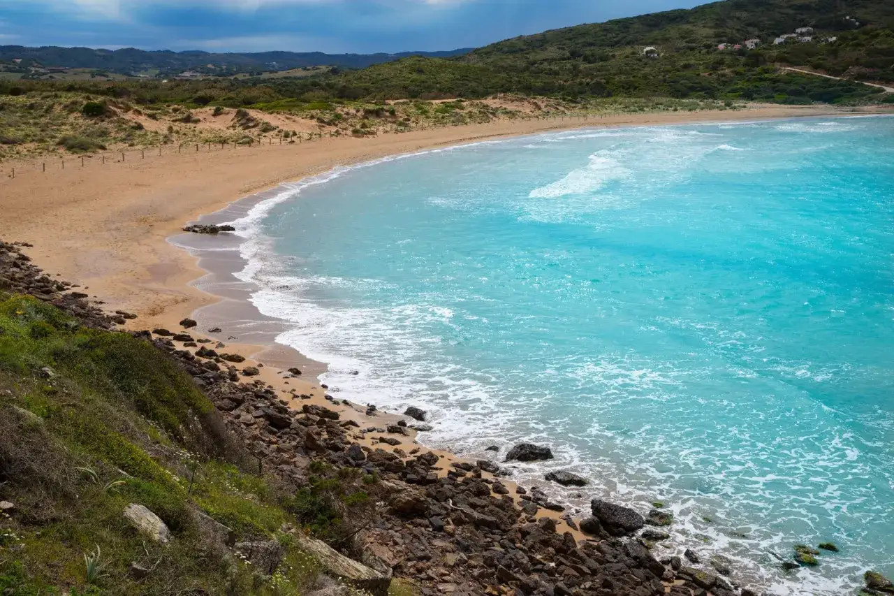 Mar turquesa y arena dorada en calas cerca de Fornells, con colinas verdes y cielo nublado.