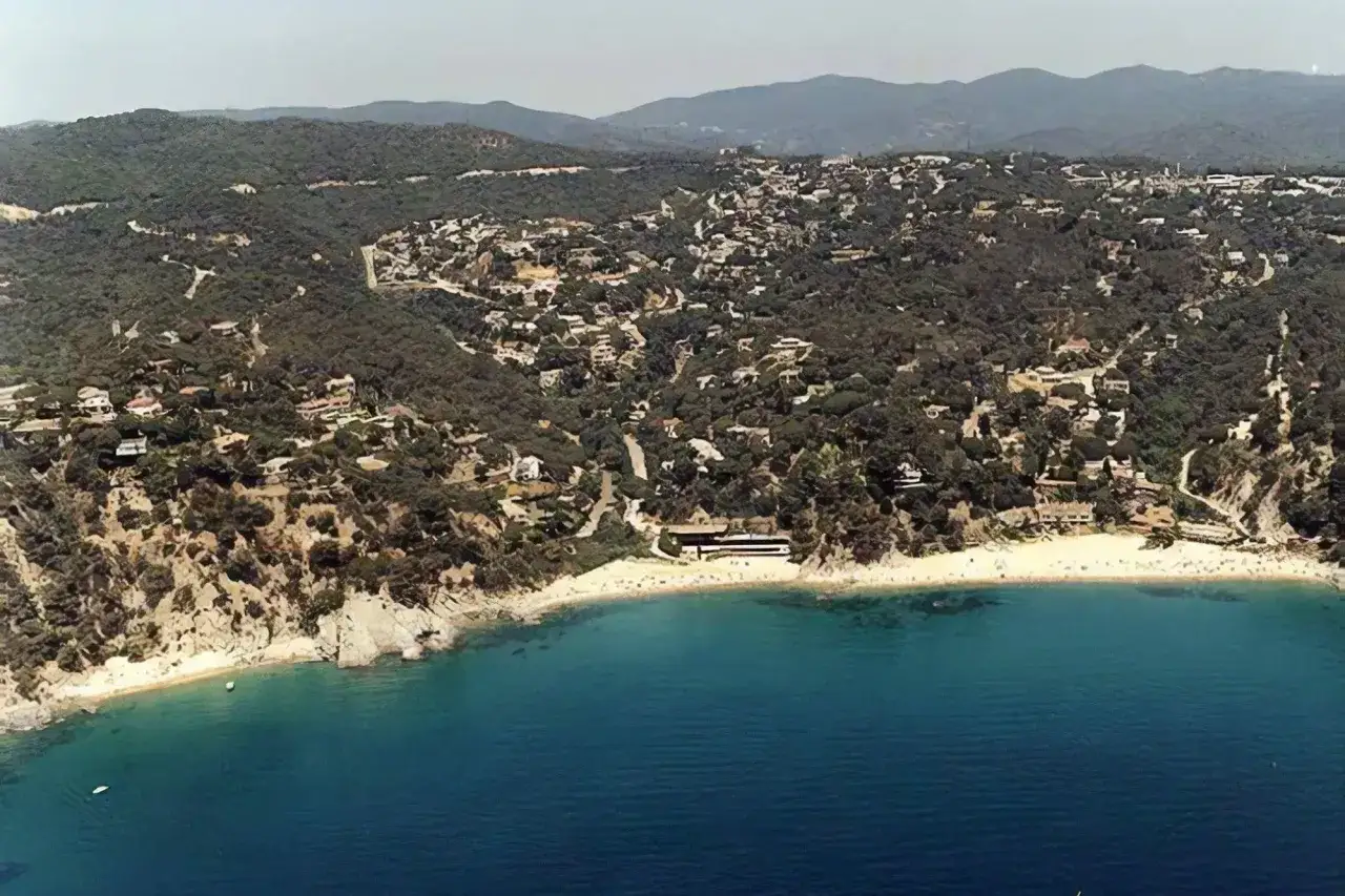 Vista aérea de la costa de Santa Maria de Llorell, con casas dispersas entre pinos y una playa dorada bañada por aguas turquesas.