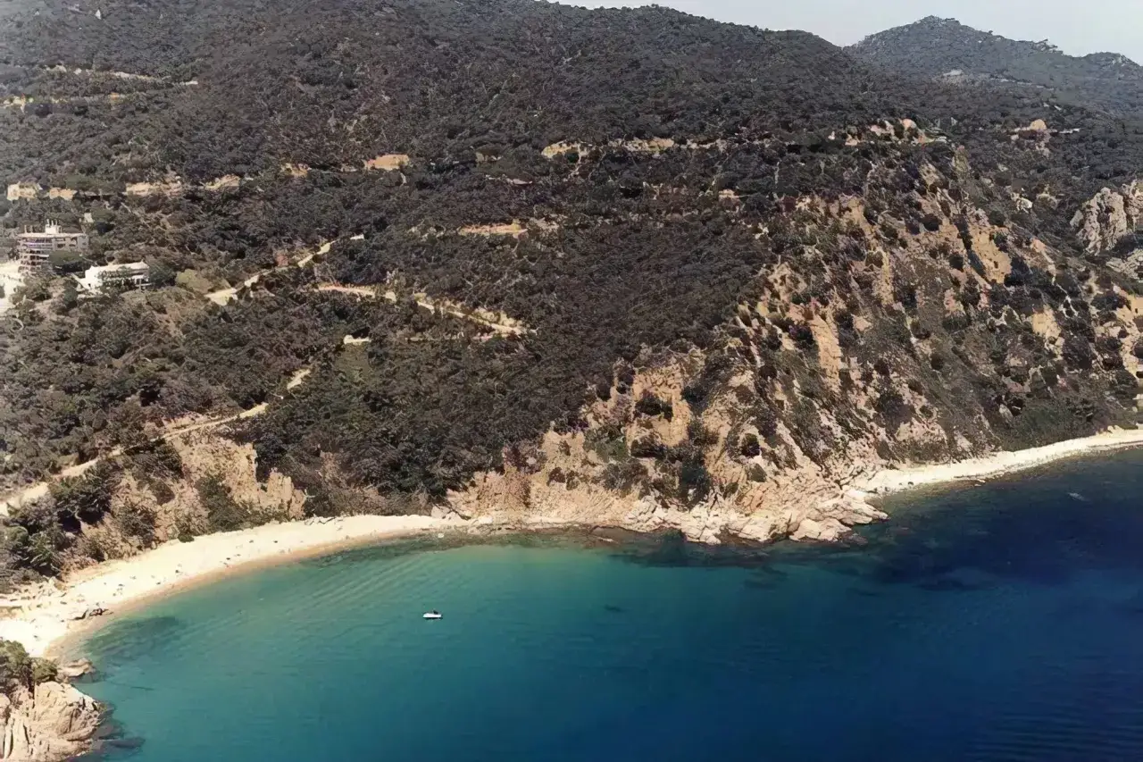 Vista a&eacute;rea de la platja de Canyerets, con arena dorada, aguas turquesas y una colina boscosa que desciende hasta el mar.