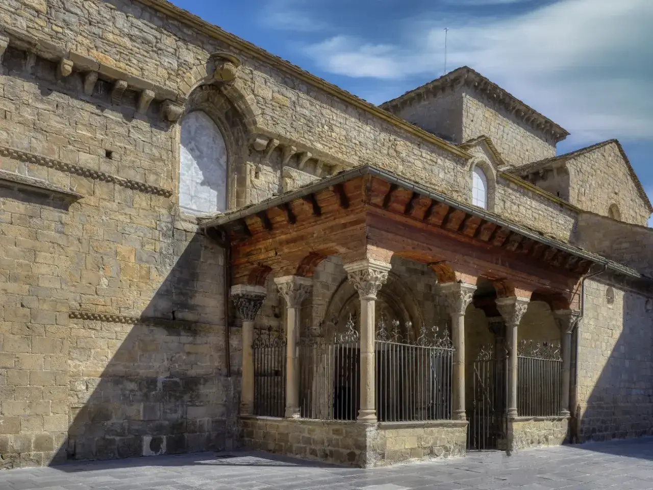 La catedral de Jaca, con su imponente arquitectura rom&aacute;nica, se alza bajo un cielo azul. Un p&oacute;rtico de madera y piedra protege la entrada.