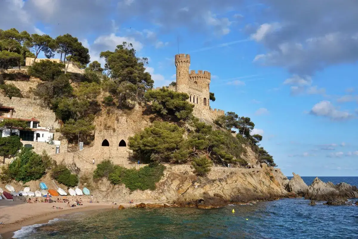 Castillo medieval sobre acantilados rocosos en sa caleta Lloret de Mar. Playa con barcas y gente disfrutando del sol.