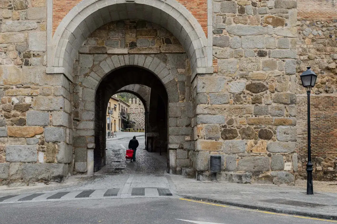Un hombre con un carrito rojo cruza un arco de piedra en Toledo. Descubre qué ver en Toledo y su historia.