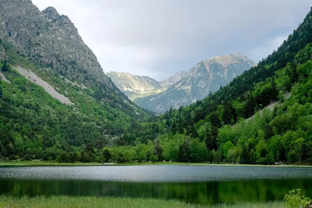 Paisaje sereno de los Lagos de Colomers, ideal para una ruta corta. Montañas verdes y un lago tranquilo reflejan el cielo nublado.