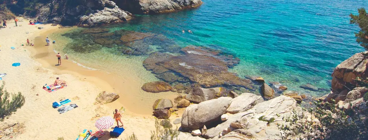 Gente disfruta del sol y el mar turquesa en la cala del pi, con rocas y arena dorada.