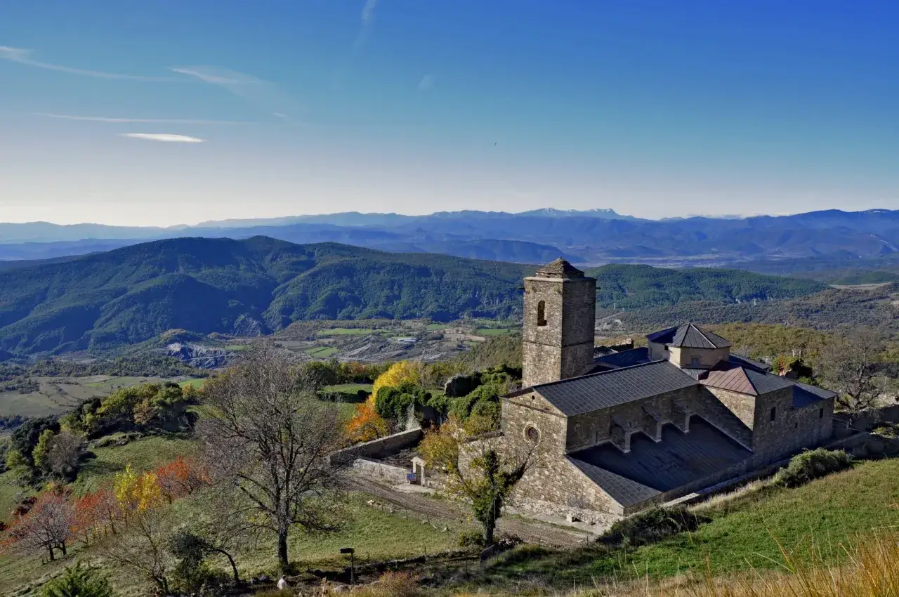 El monasterio de San Victorián se alza majestuoso entre montañas verdes y un cielo azul intenso, con árboles de otoño en primer plano.