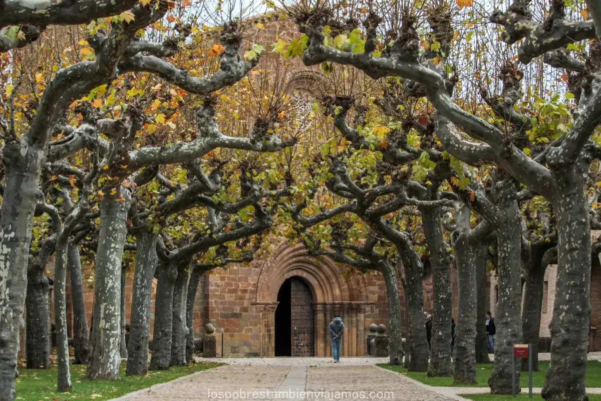 Un camino flanqueado por árboles nudosos conduce a la entrada románica del monasterio de Veruela. Las hojas otoñales añaden color a la escena.