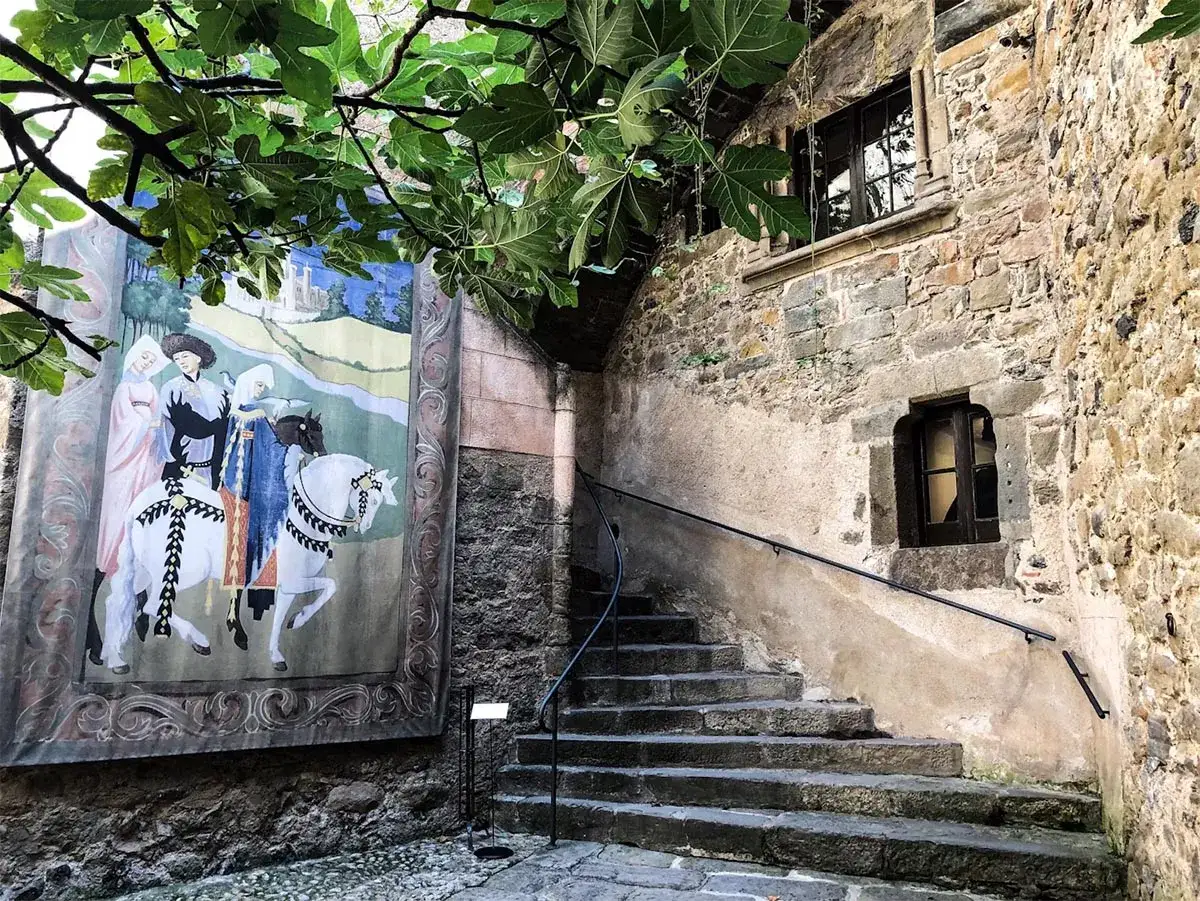 Un tapiz con figuras a caballo decora la pared de un patio empedrado junto a una escalera de piedra. El tapiz evoca la grandeza de un castell de gala.