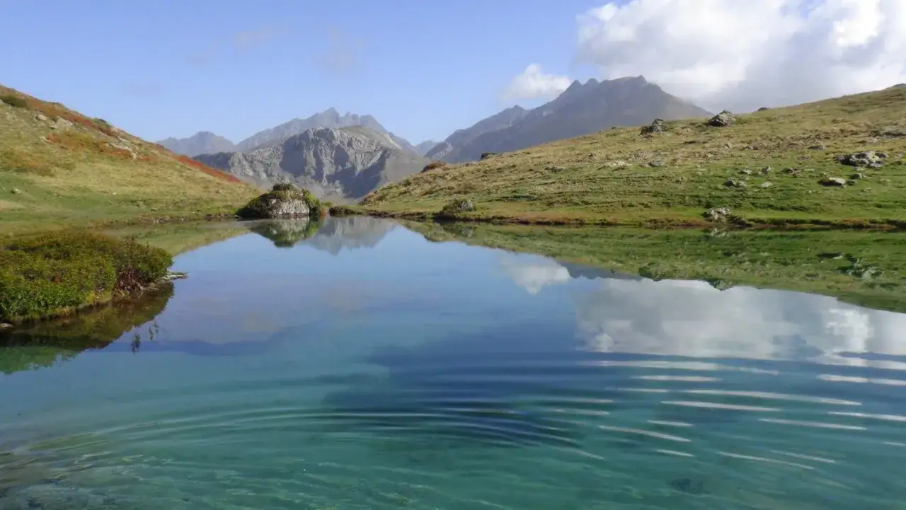 Un sereno ibón de espelunciecha refleja montañas y un cielo azul con nubes.