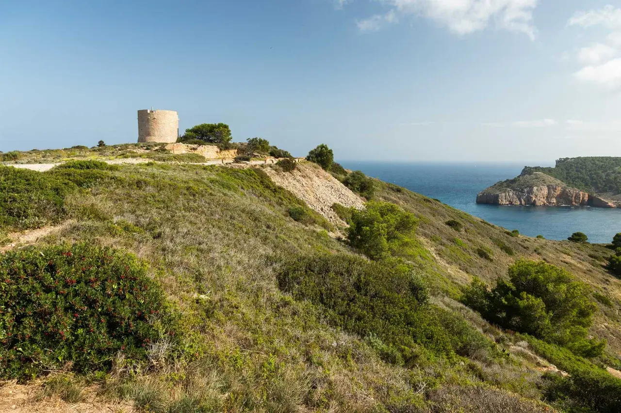 La torre de Montg&oacute; se alza sobre una colina verde, vigilando el mar azul y una bah&iacute;a rocosa.