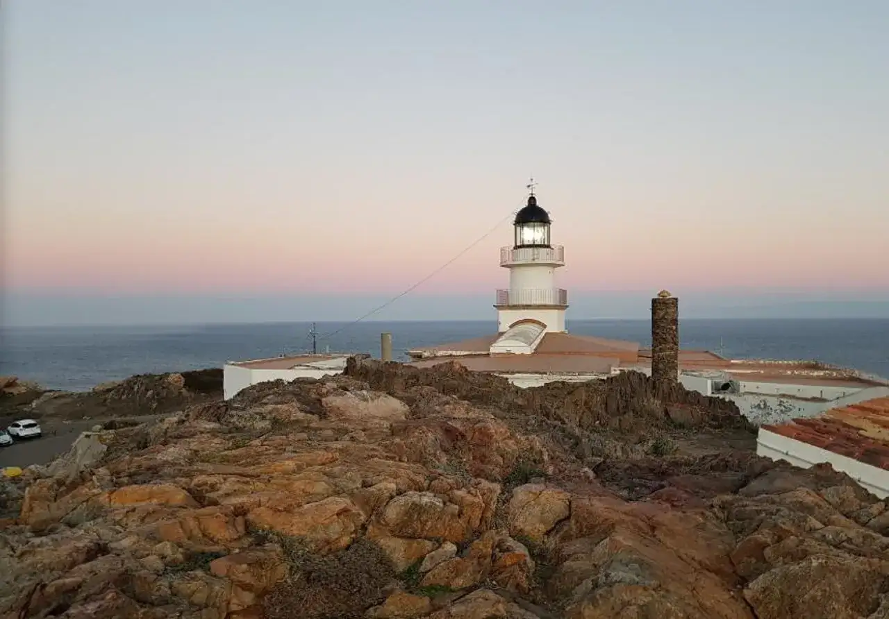 Faro Cap de Creus al atardecer, con el mar y el cielo en tonos rosados. Rocas rojizas en primer plano.
