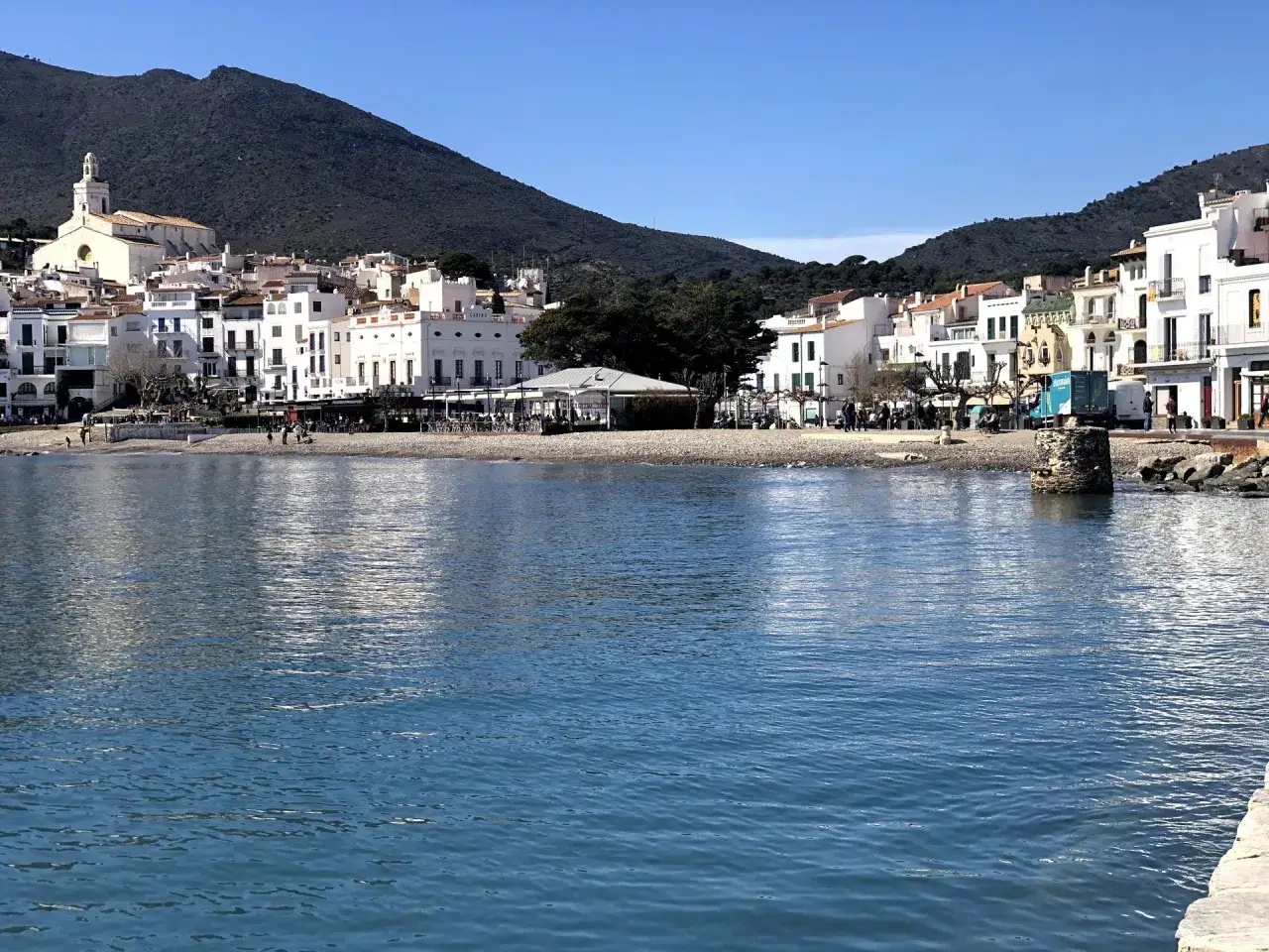 Vista del mar azul frente a la platja gran Cadaqués, con casas blancas y una iglesia en la ladera de una montaña verde.