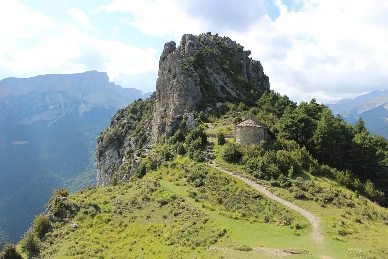 Pequeña ermita de piedra en el valle de Chistau, rodeada de vegetación y con imponentes montañas al fondo.