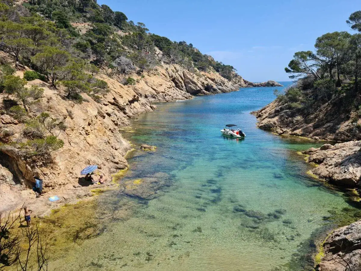 Aguas cristalinas en las calas de Tossa de Mar. Una peque&ntilde;a barca descansa en la bah&iacute;a, mientras unas personas disfrutan del sol en la orilla rocosa.