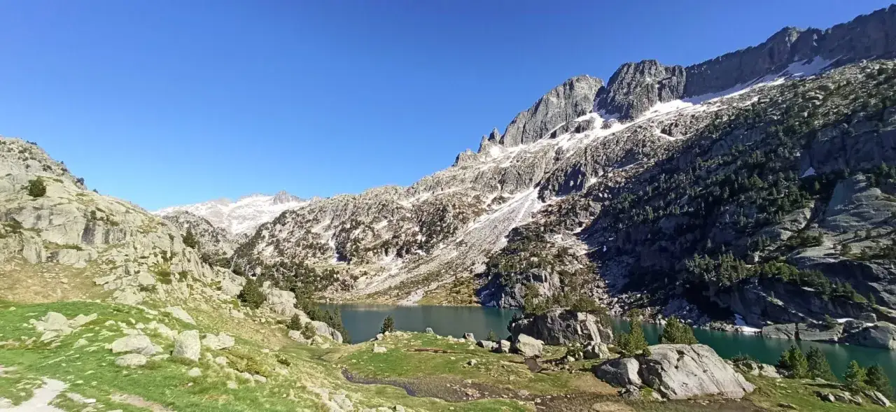 Lago sereno en el Vall de Bo&iacute;, rodeado de rocas, prados verdes y monta&ntilde;as nevadas bajo un cielo azul intenso.